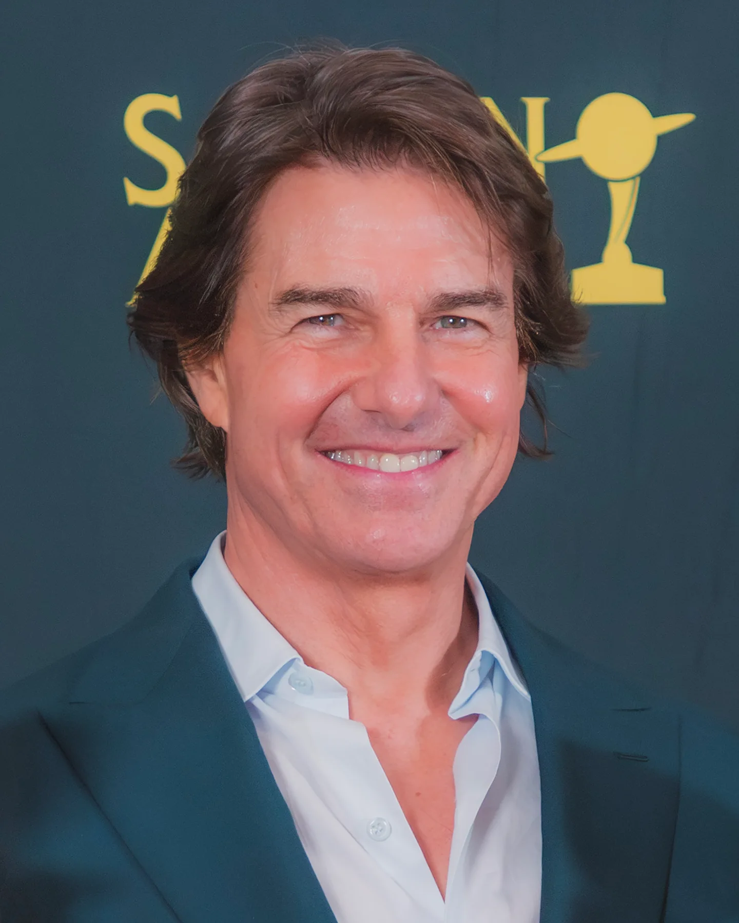 Tom Cruise smiling in a dark green jacket and open-collared light blue shirt at an awards event
