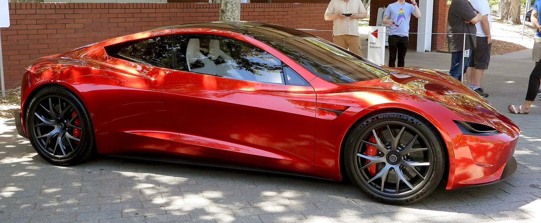 Red Tesla Roadster prototype parked on a brick-paved street, side view, as visitors look on