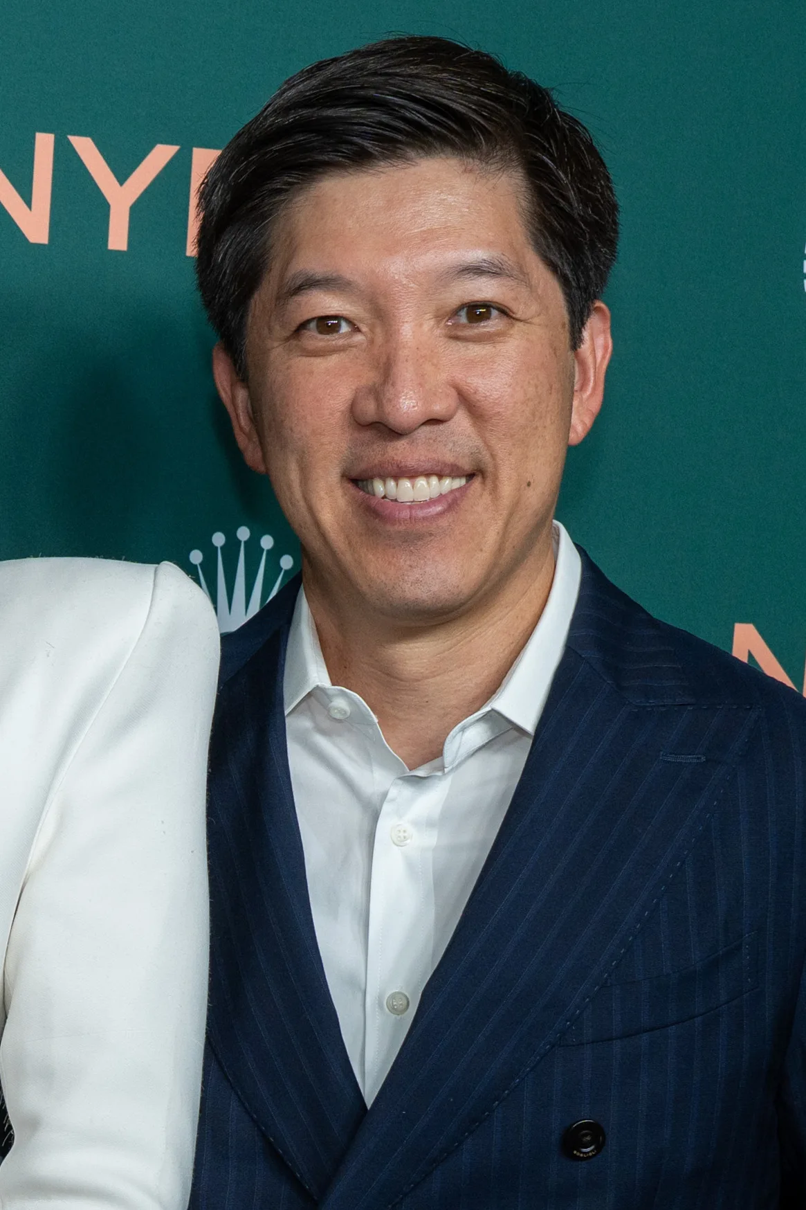 Dan Lin smiling in a navy pinstripe suit and white shirt in front of a green event backdrop