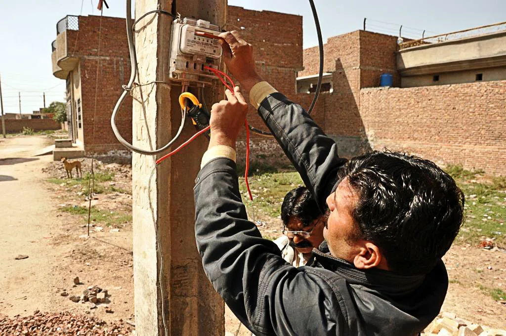 Technician tests a household electricity meter on a concrete pole beside brick houses using a clamp meter