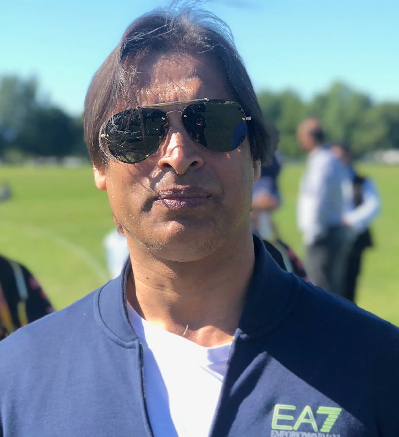 Shoaib Akhtar in aviator sunglasses and a navy jacket at a sunny cricket ground