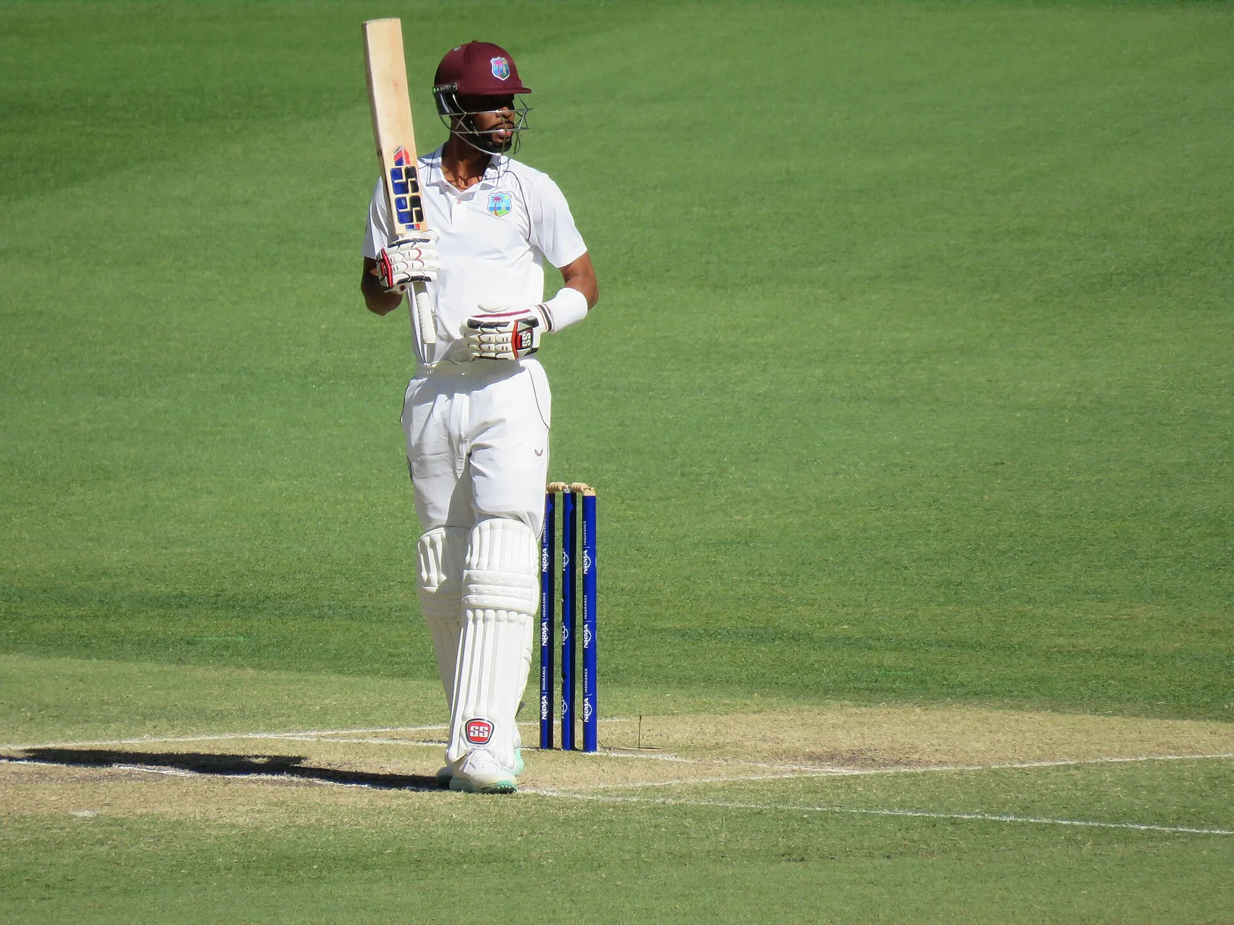 West Indies batter in Test whites raising his bat beside the stumps on a green outfield