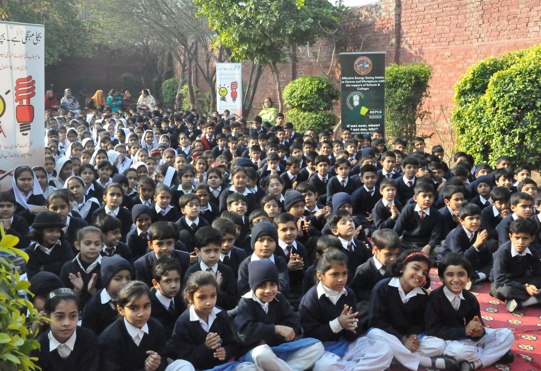Rows of schoolchildren in navy uniforms and white shirts seated on the ground at an outdoor school assembly