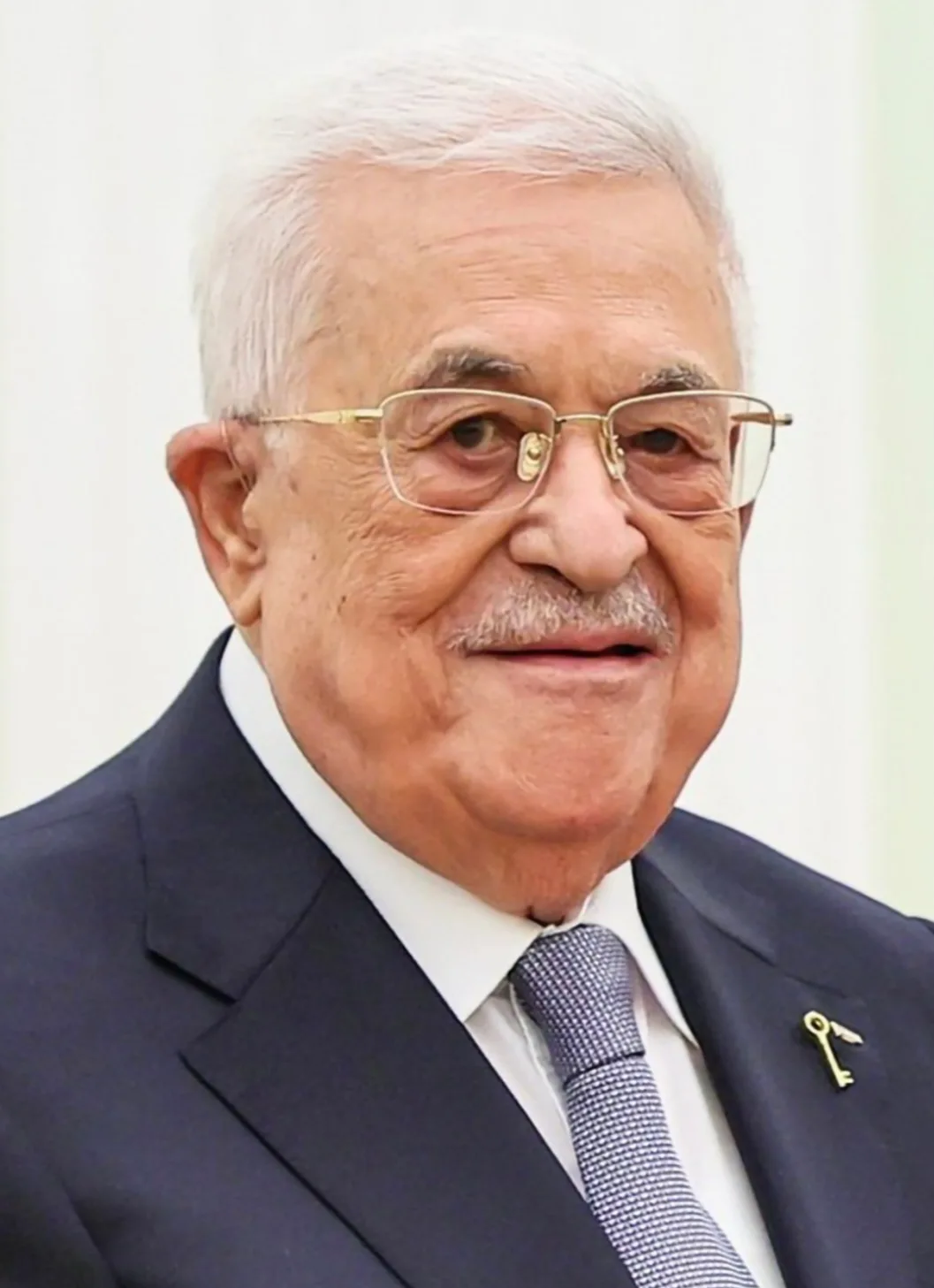 Mahmoud Abbas in a dark suit and glasses, smiling, with a small key pin on his lapel