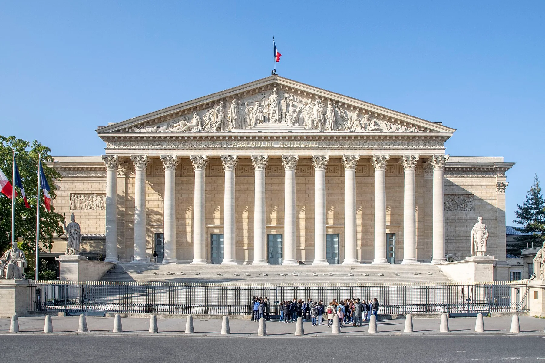 The columned front of France's National Assembly building with a French flag on top and a group of visitors at the gate