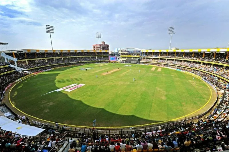 Packed stands around a green oval cricket ground with floodlight towers during a match in Indore