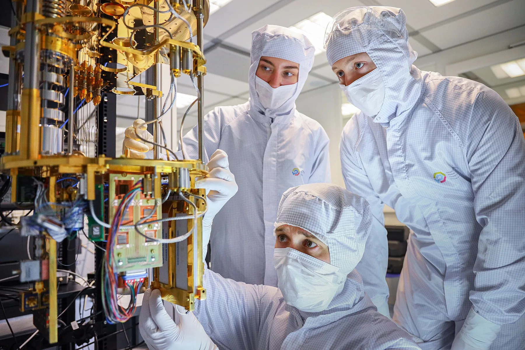 Three researchers in white cleanroom suits inspect the gold-coloured wiring of a quantum computer in a laboratory