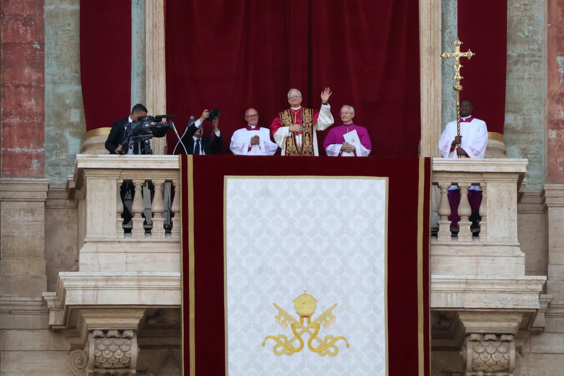 Pope Leo XIV waves from the balcony of St Peter's Basilica, flanked by clergy and a cameraman