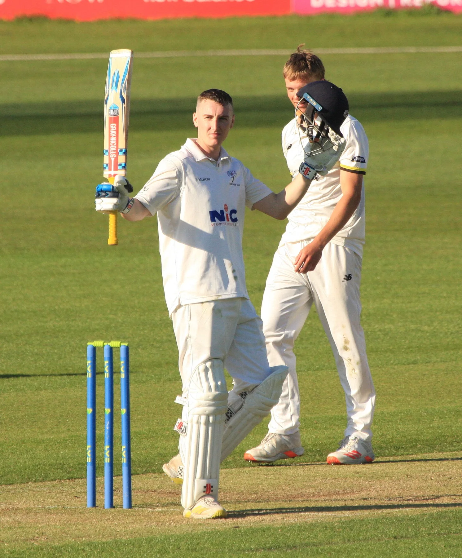 Harry Brook raises his bat in Yorkshire whites after a century as a teammate in a helmet congratulates him