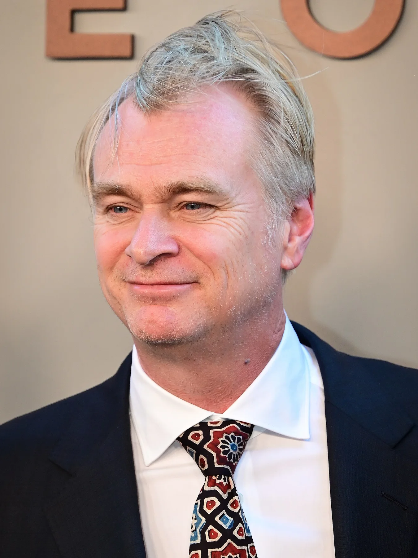 Christopher Nolan smiling at a premiere, in a dark suit and patterned red and blue tie