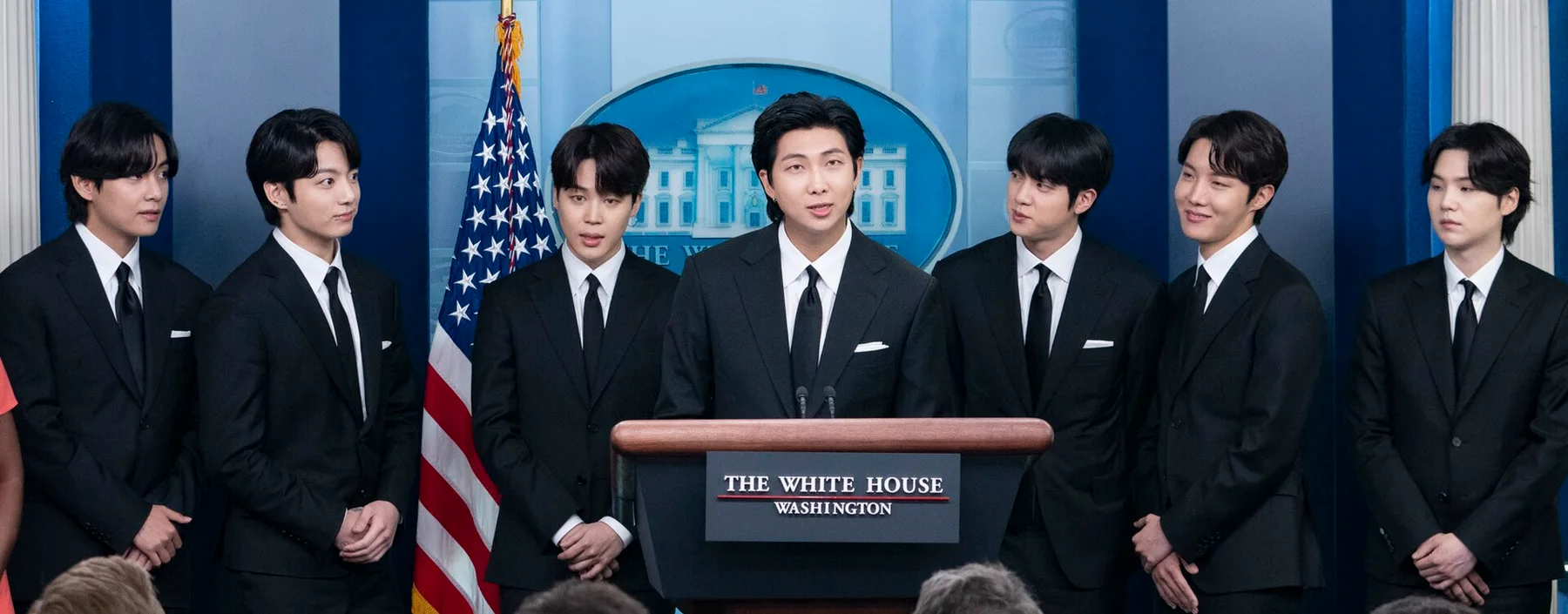 The seven members of BTS in dark suits standing behind a podium at a White House briefing