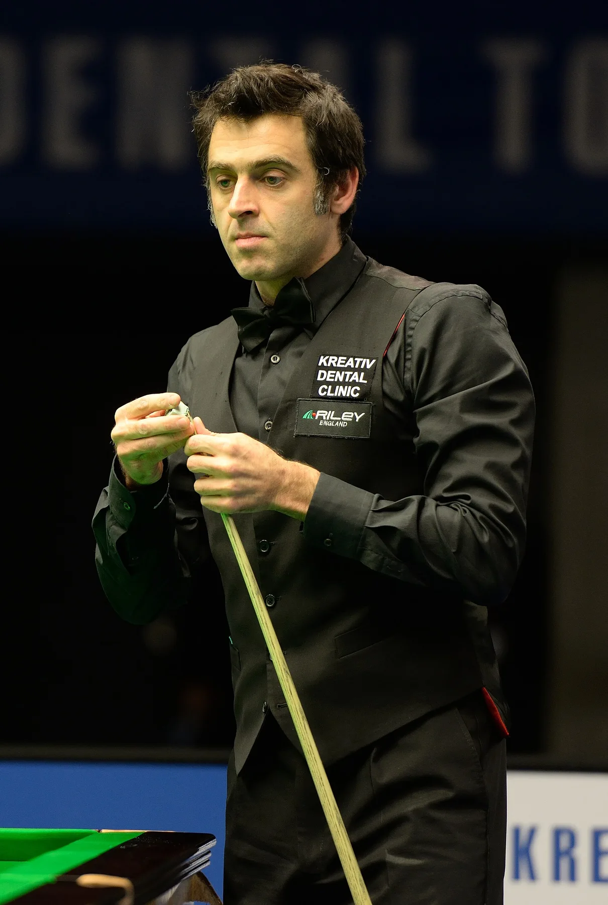 Ronnie O'Sullivan in waistcoat and bow tie chalks his cue beside a snooker table