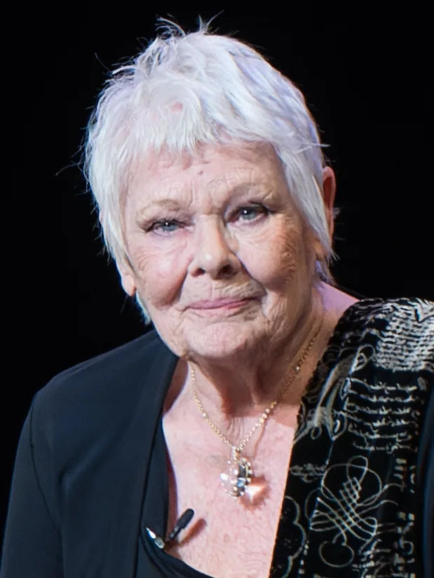 Judi Dench, with short white hair and a pendant necklace, smiles against a dark background