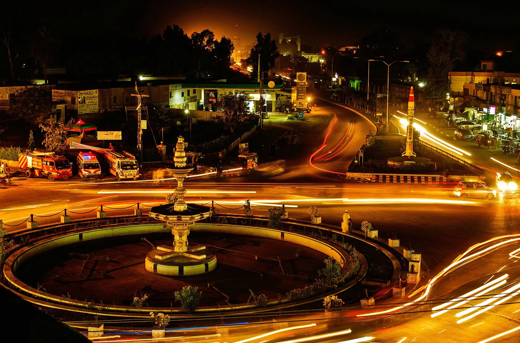 Night view of a lit roundabout fountain in Hafizabad, with Rescue 1122 vehicles parked by the road
