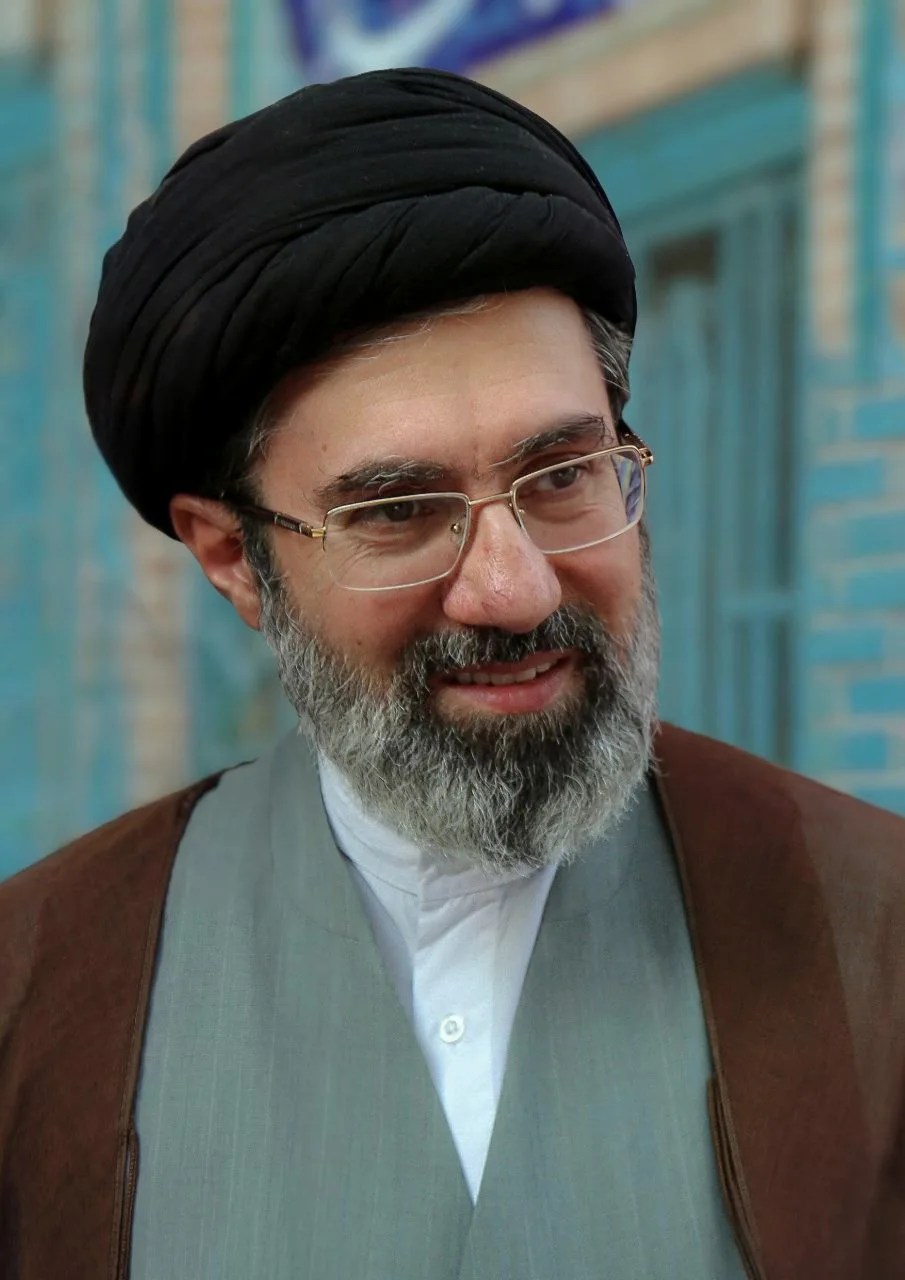 Mojtaba Khamenei in a black turban and glasses, smiling, in front of blue tiled wall