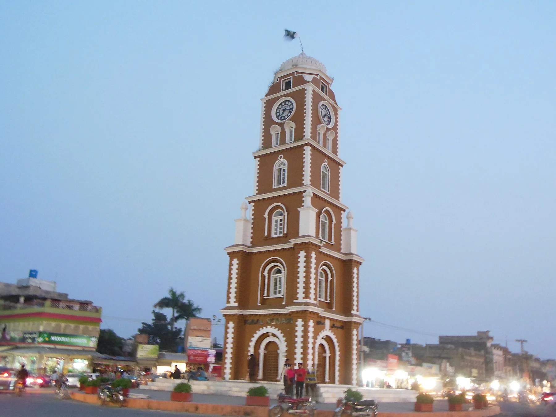 Sialkot clock tower at dusk with motorcycles and shops around the roundabout