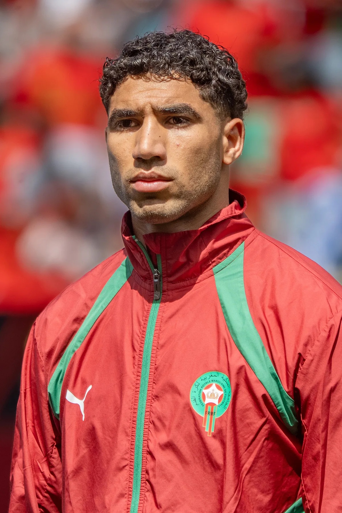 Achraf Hakimi in a red and green Morocco team jacket before a match, crowd blurred behind