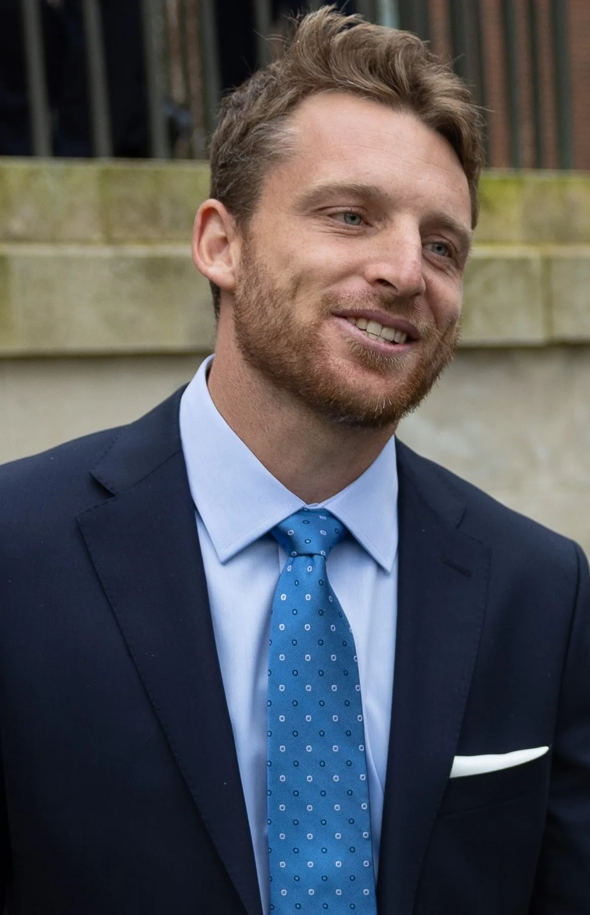 Jos Buttler smiling in a navy suit, light blue shirt and spotted blue tie outdoors