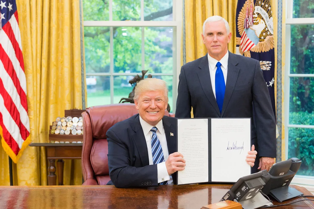 Donald Trump smiling at the Oval Office desk, holding up a signed executive document