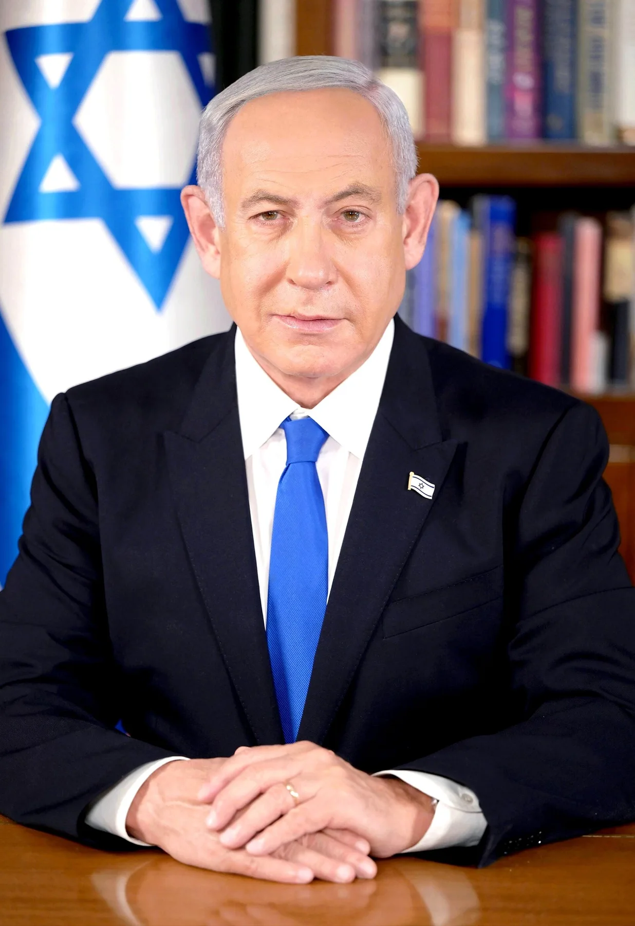 Benjamin Netanyahu in a dark suit and blue tie seated at a desk before an Israeli flag and bookshelves