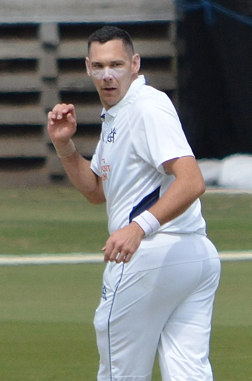 Scott Boland in white cricket kit on the field, gesturing with his right hand