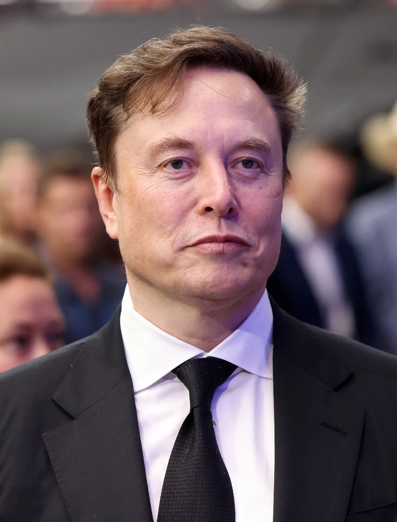 Elon Musk in a dark suit and black tie at an indoor event, with a crowd behind him
