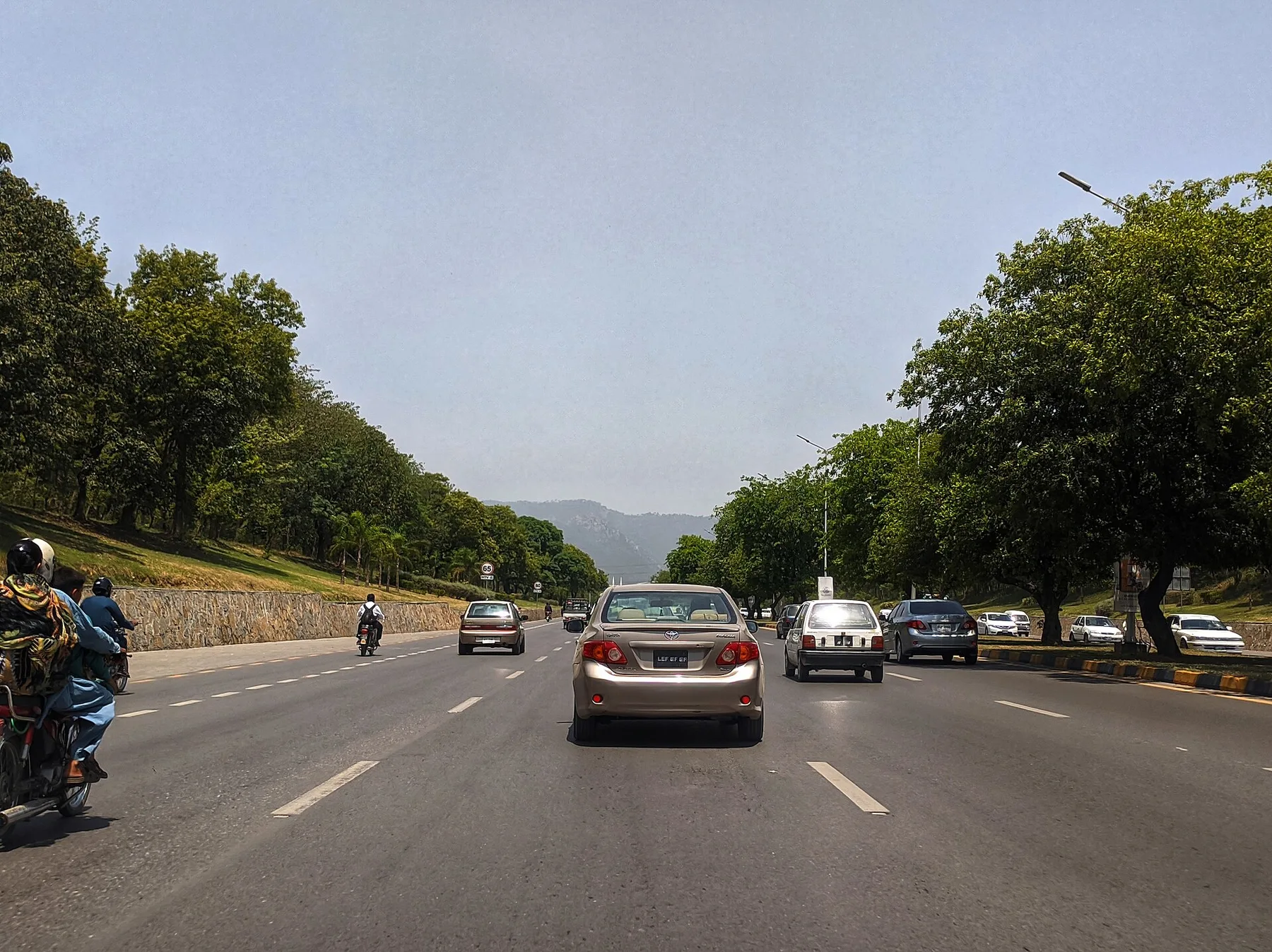 Cars and motorcycles on a wide tree-lined Islamabad road with green belts and hills ahead