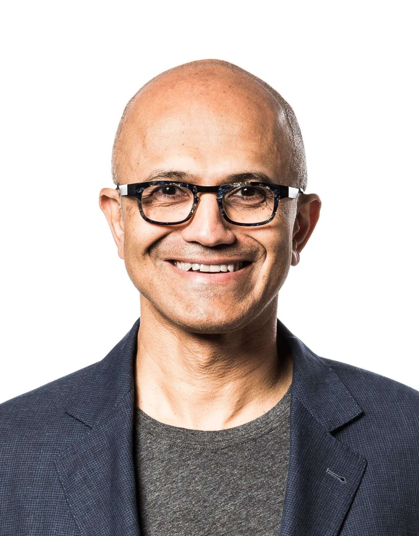 Satya Nadella smiling in glasses, dark jacket and grey T-shirt against a white background