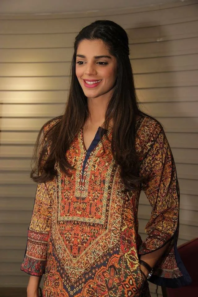 Sanam Saeed smiling in an orange and blue printed kurta with long dark hair, standing indoors
