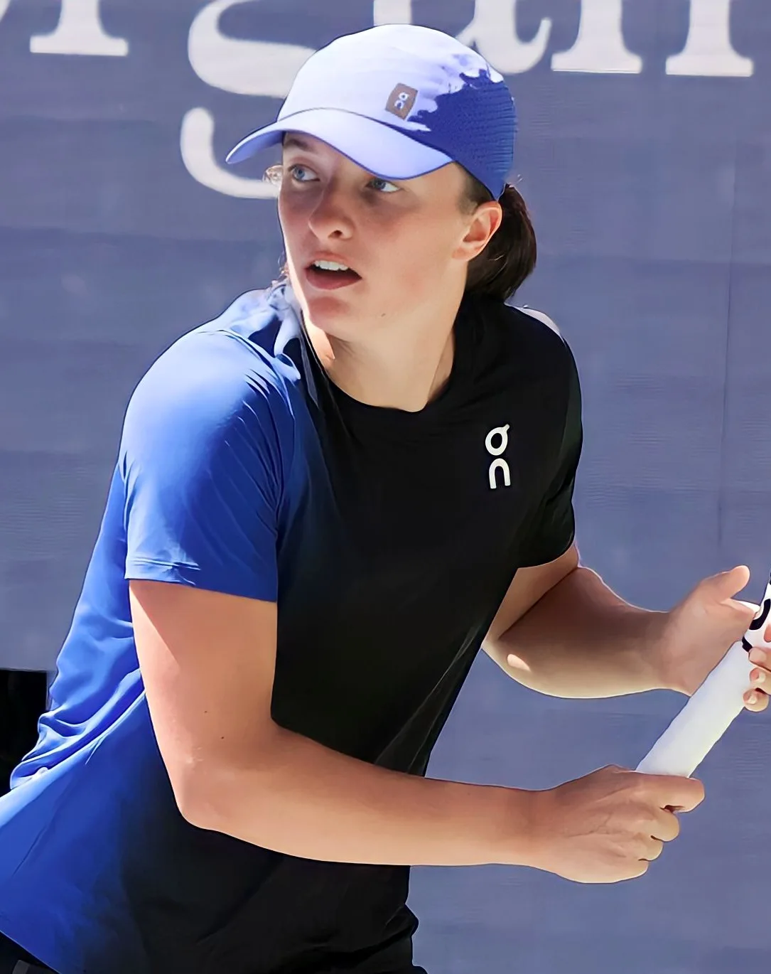 Iga Swiatek in a white cap and dark shirt holding her racket during a match on a blue court