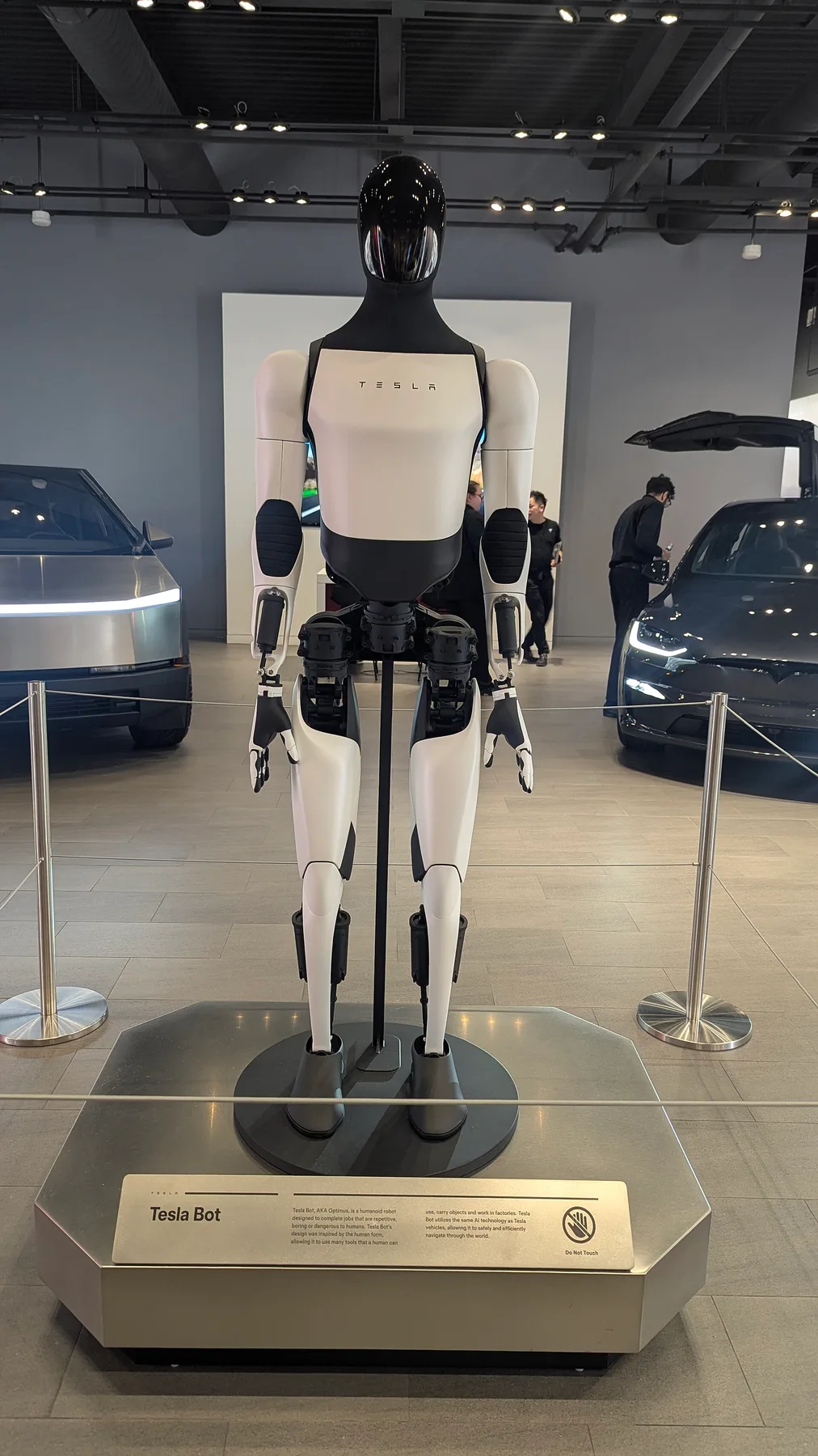 Tesla Optimus humanoid robot on a display stand in a showroom, with a Cybertruck and Model X behind