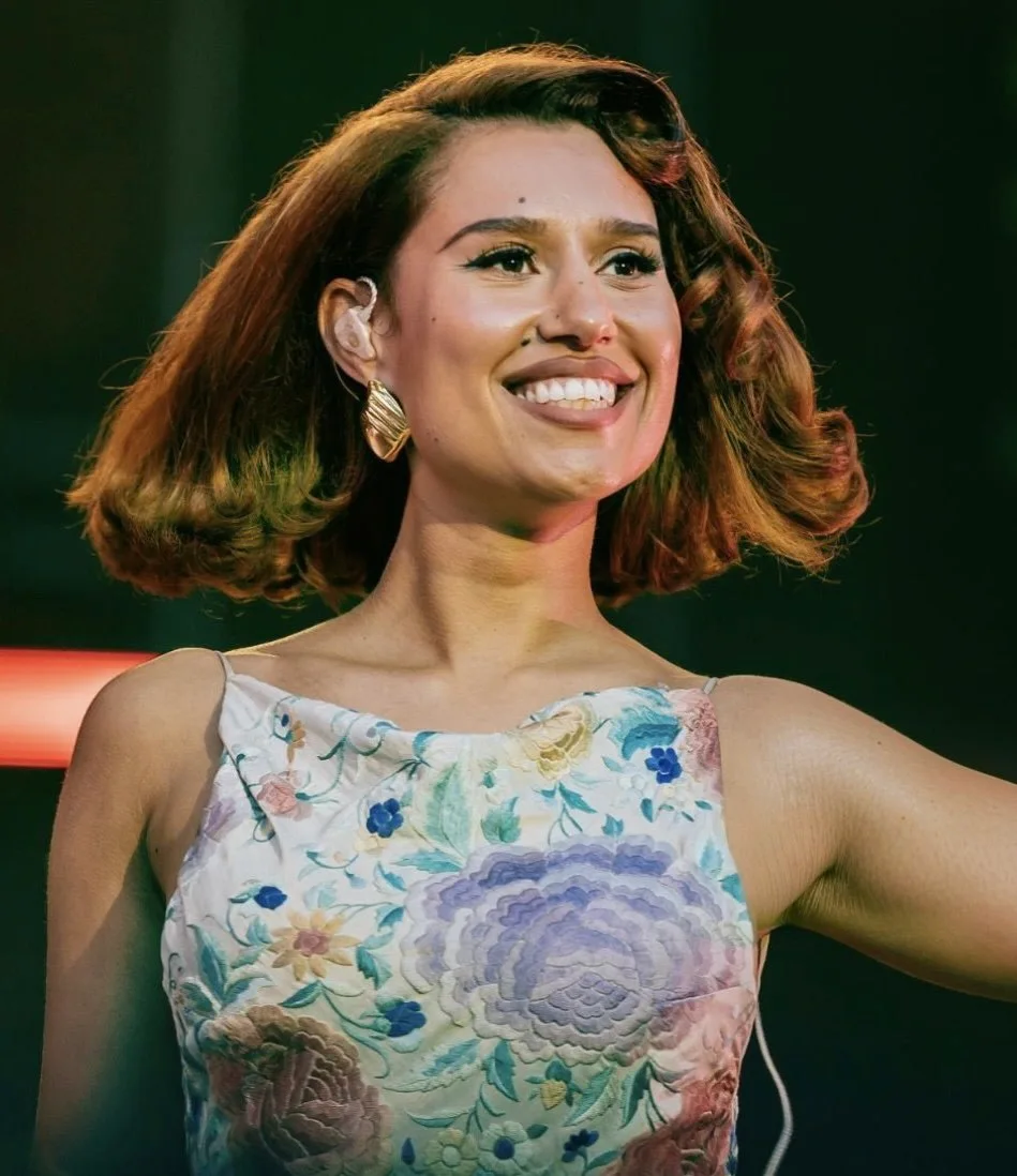 Raye smiling on stage in a floral embroidered dress, one arm stretched out towards the crowd