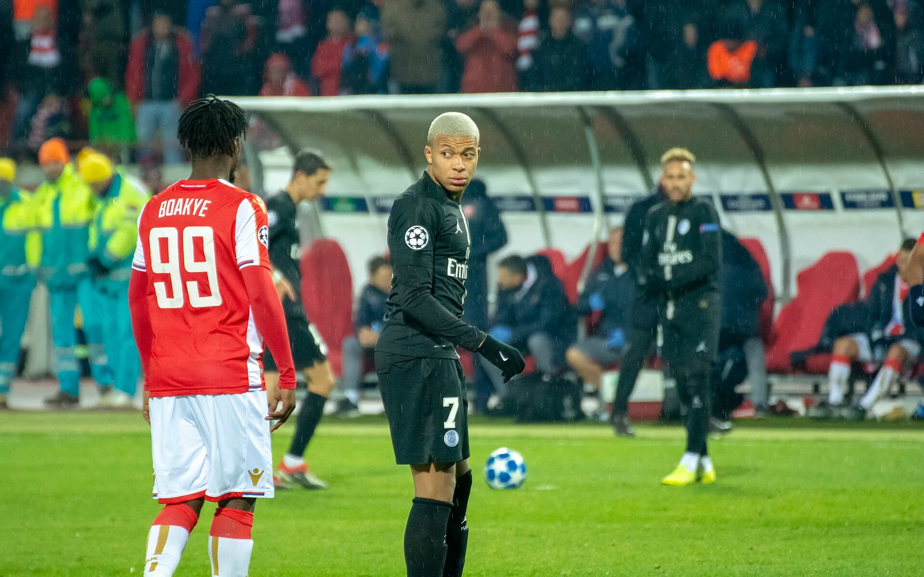 Kylian Mbappe in a black PSG number 7 kit on the pitch beside a player in red, number 99