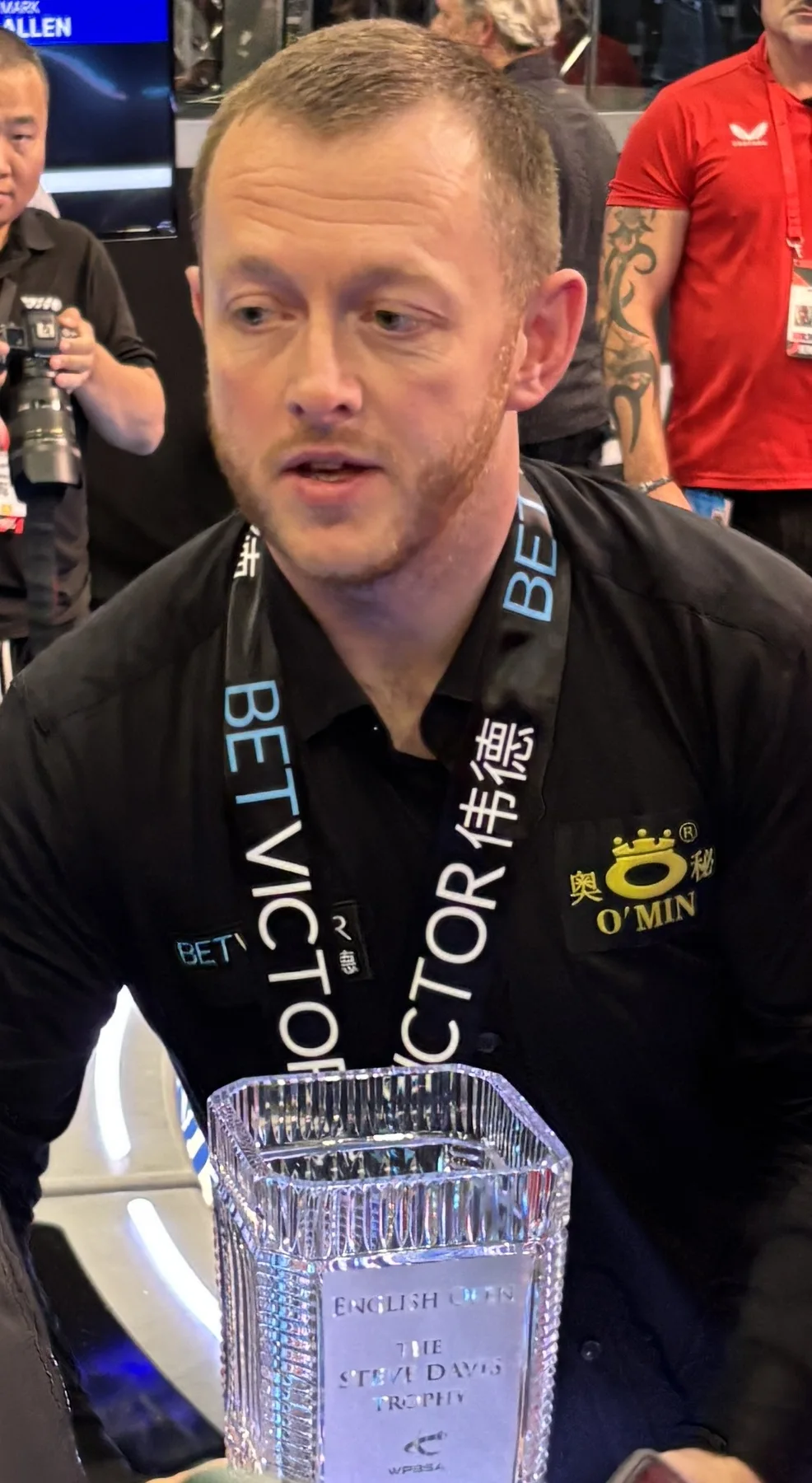 Mark Allen in a black sponsor shirt speaks to reporters beside the English Open crystal trophy