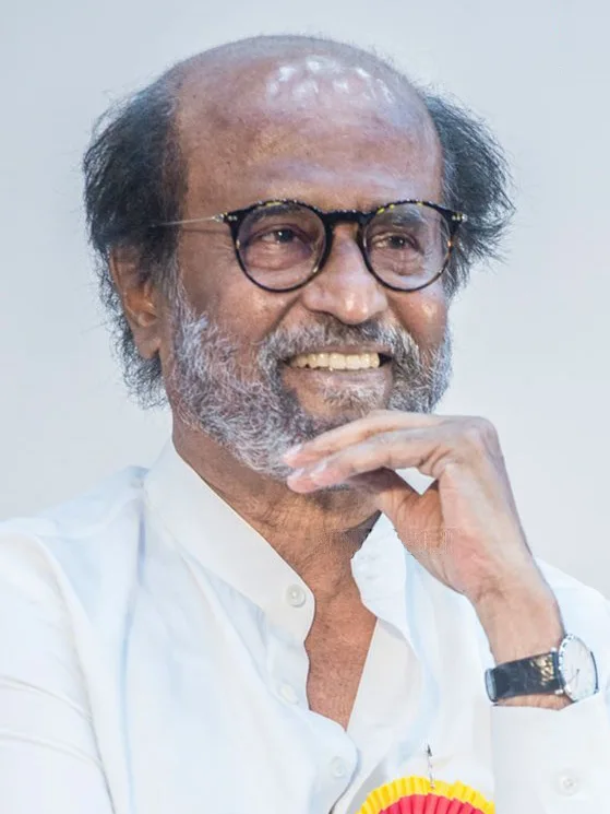 Rajinikanth in round glasses and a white shirt, smiling with his hand under his chin
