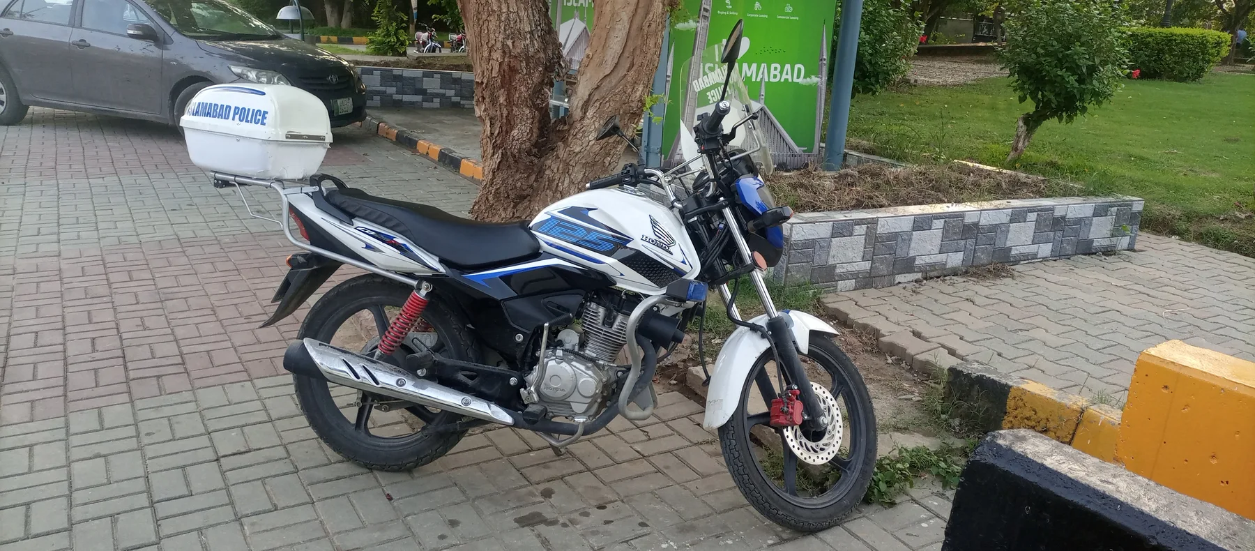 White Honda motorcycle with an Islamabad Police box parked on a paved walkway beside a tree