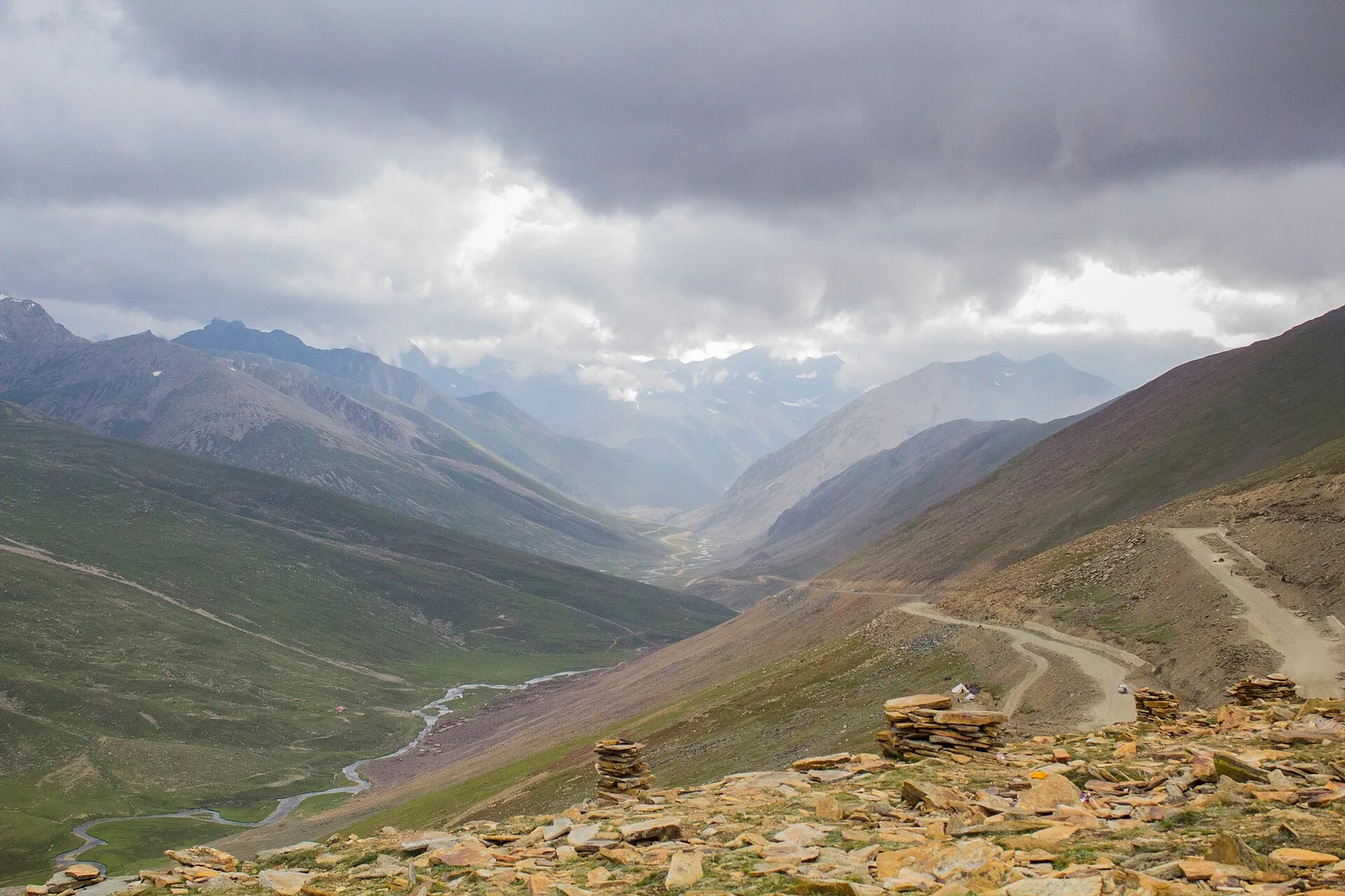 Winding Babusar Pass road through mountain valley under heavy grey clouds, with stone cairns in the foreground