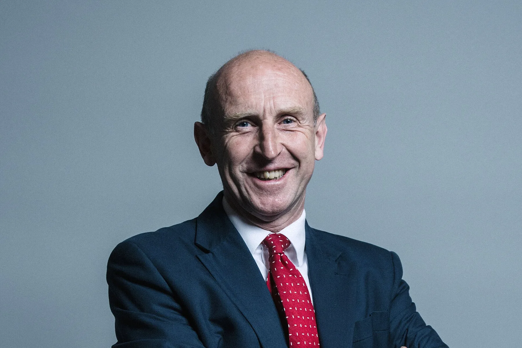 John Healey smiling in a navy suit and red patterned tie against a plain grey background