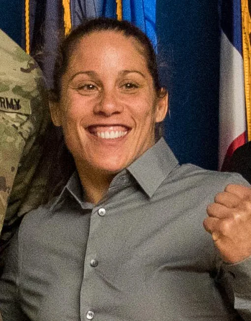 Liz Carmouche smiling and raising a fist in a grey shirt, with flags behind her