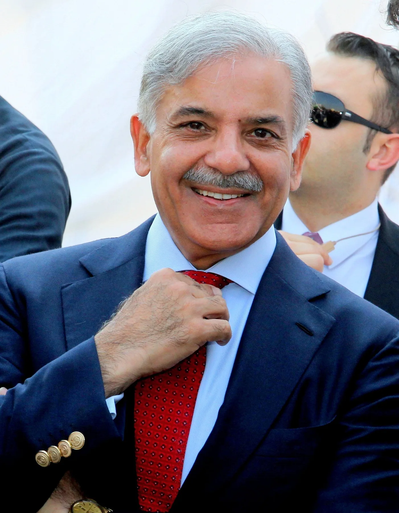 Shehbaz Sharif smiling and adjusting a red tie, wearing a navy blazer outdoors