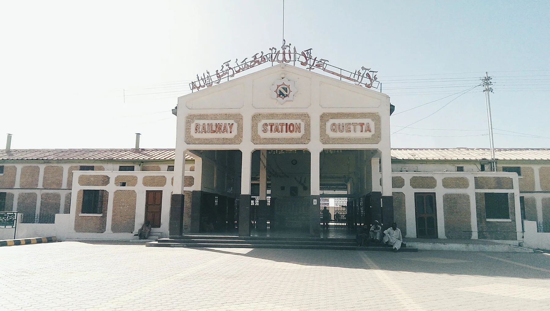 Front entrance of Quetta Railway Station with its name in red letters and men seated on the steps