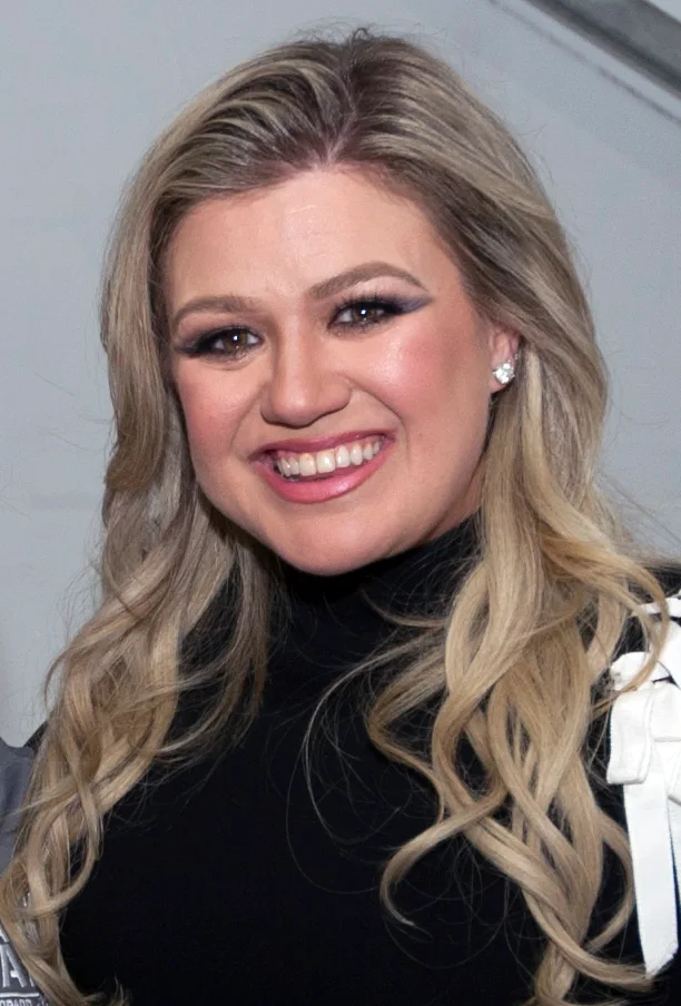 Kelly Clarkson smiling at an event, with long wavy blonde hair and a black top