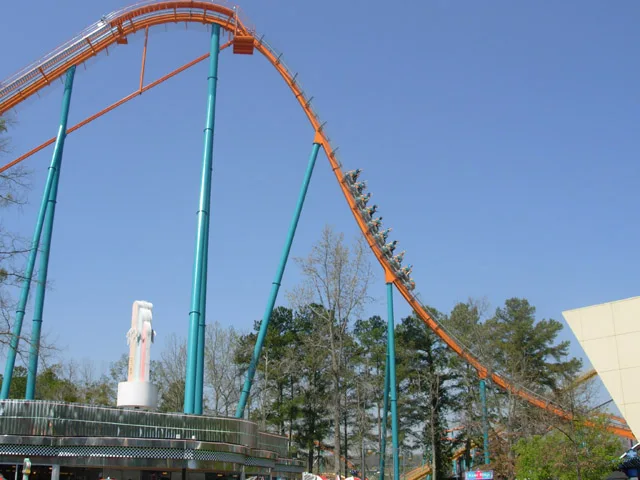 Riders on a train descending the orange track of a tall roller coaster with teal supports under a blue sky