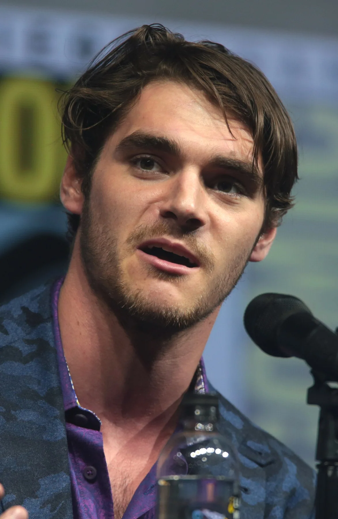 RJ Mitte speaking into a microphone on a convention panel, wearing a blue camouflage jacket