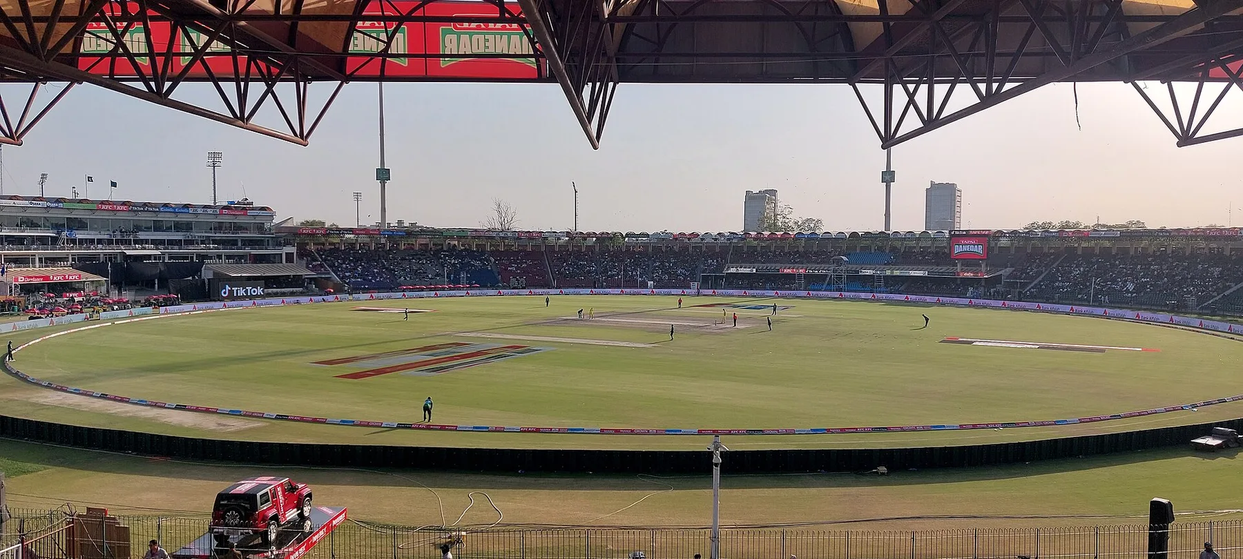 Wide view of a cricket ground in Lahore under the stand roof, with the pitch and sightscreen in the sun