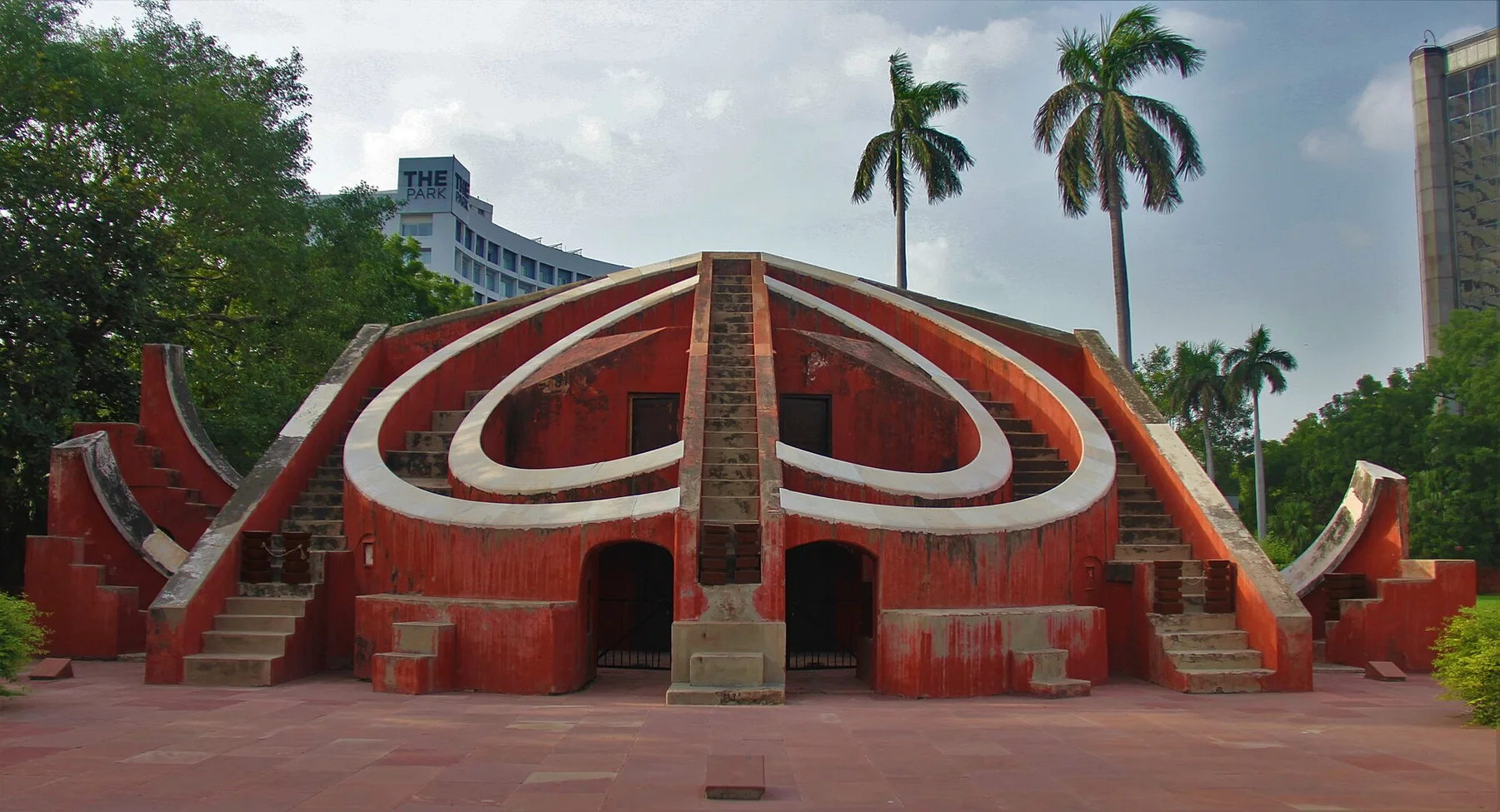 Red sandstone Mishra Yantra instrument at Jantar Mantar in New Delhi with palm trees and a hotel behind