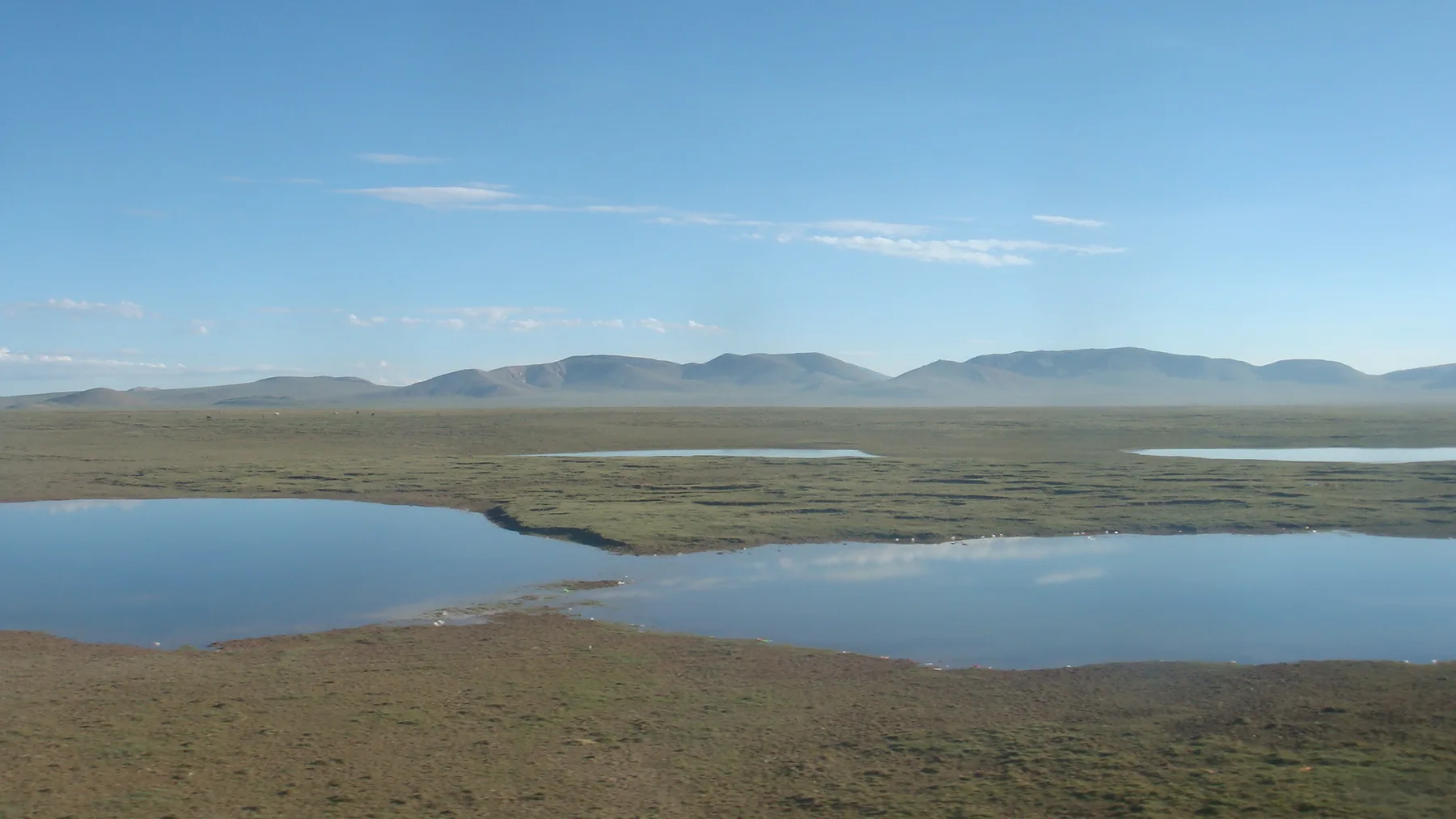 Open grassland and shallow lakes below low brown hills under a blue sky on the Tibetan plateau