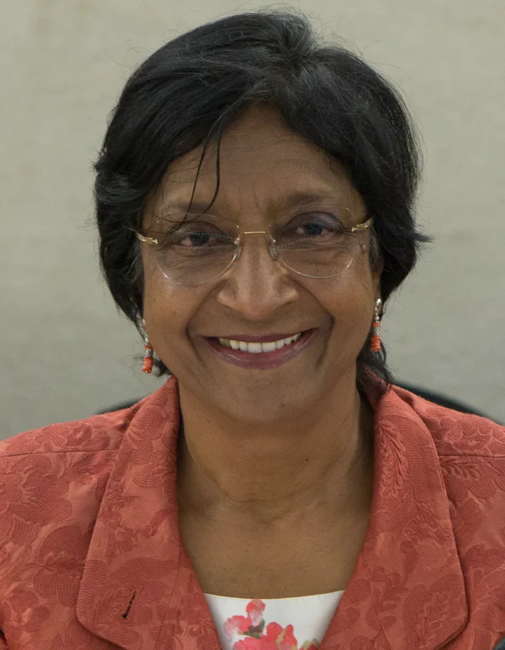 Navi Pillay smiling in rimless glasses, coral earrings and a coral patterned jacket