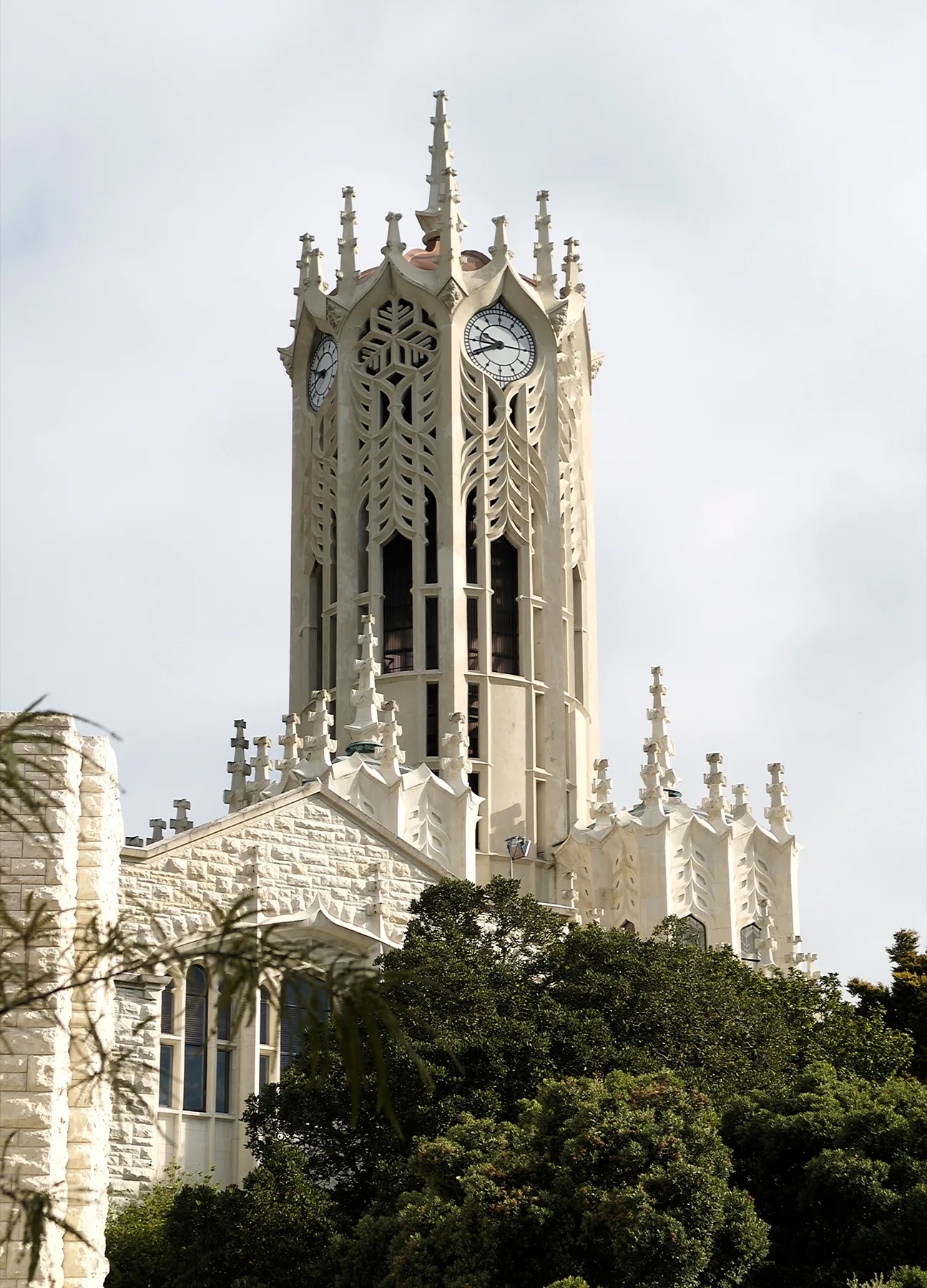 The white Gothic clock tower of the University of Auckland rising above green trees