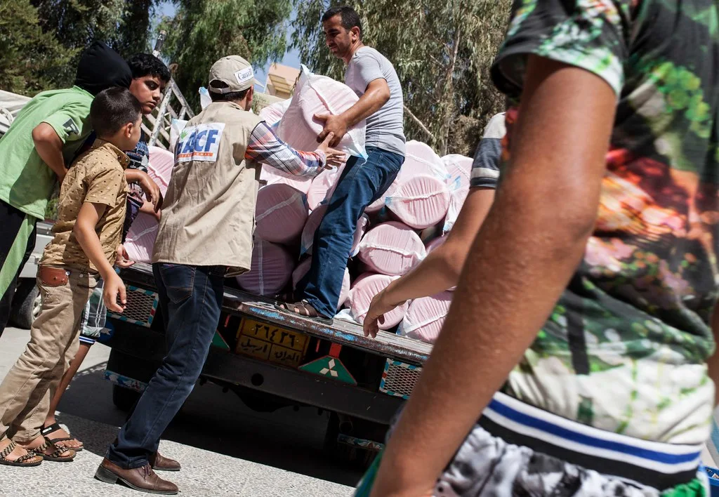 Action Against Hunger worker in a branded vest unloading aid bundles from a truck as children watch